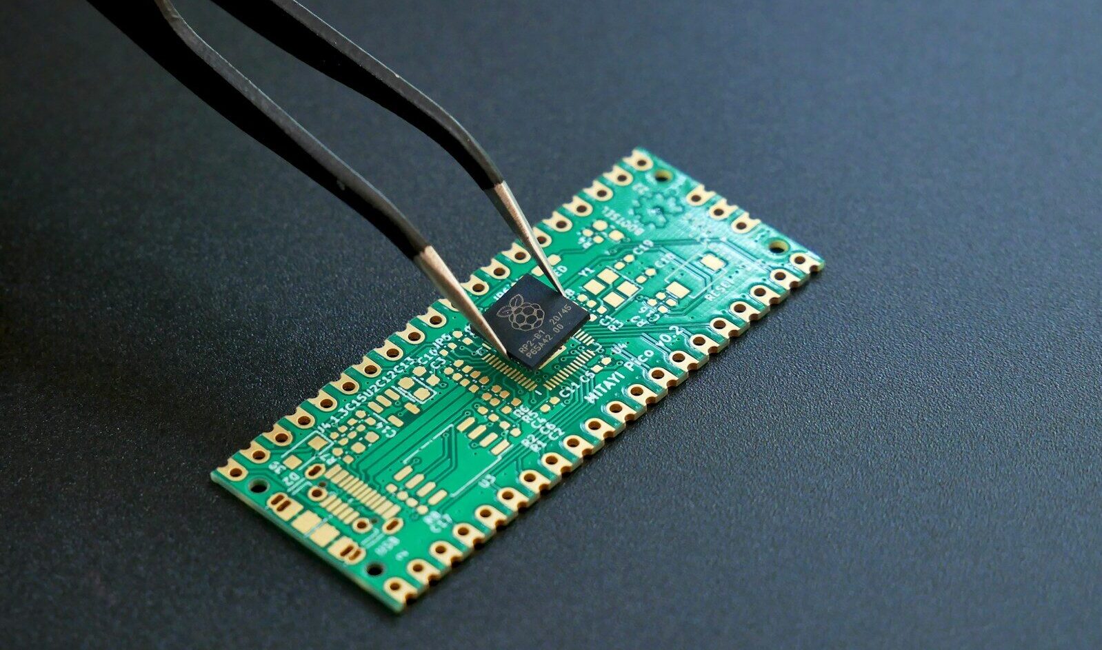Tweezers placing a small black microchip onto a green circuit board